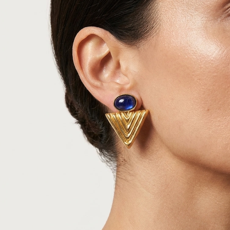 Close-up of a woman wearing gold earrings with a blue gemstone on a neutral background